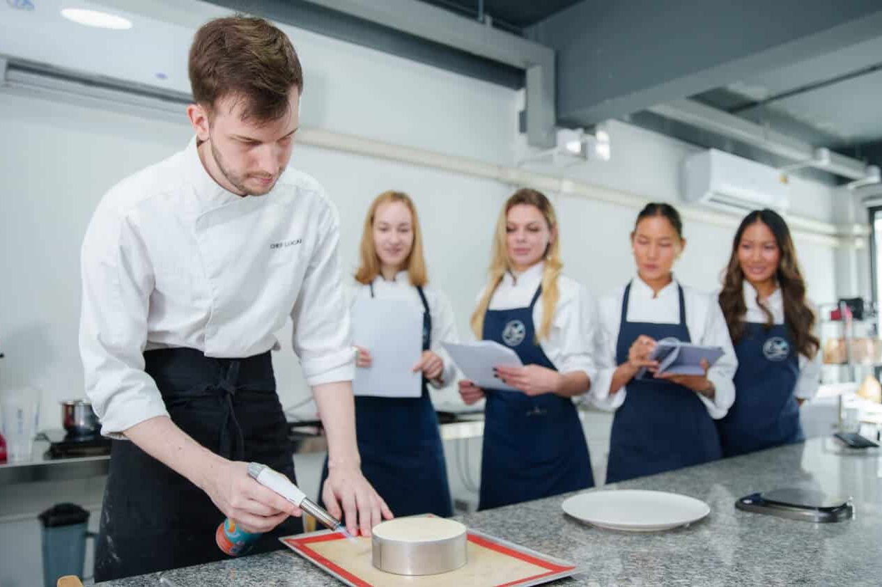 Chef Lucas demonstrates the art of making New York cheesecake while four students attentively watch in the background.