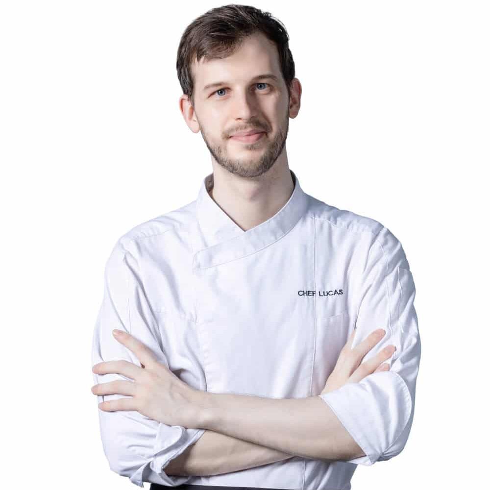 Chef Lucas wearing white chef coat and black apron standing with arm crossed in front of white background.