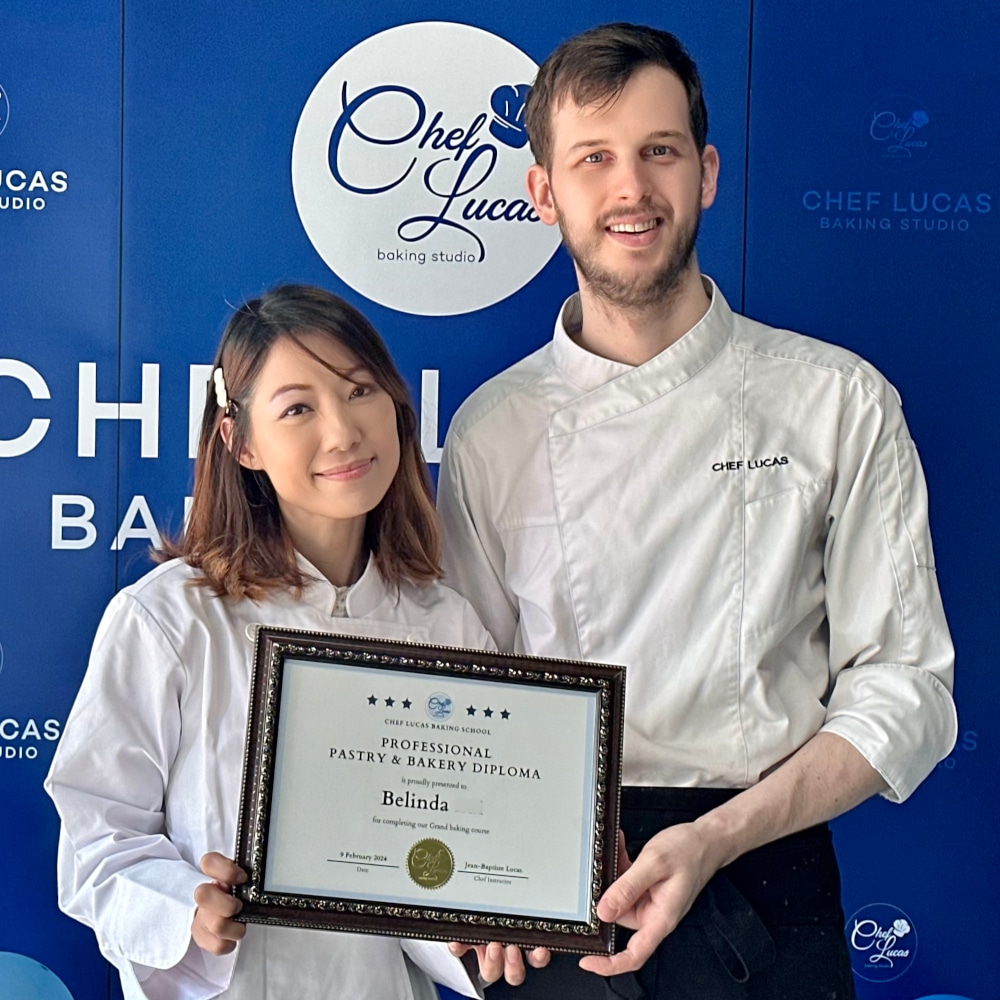 Female student and chef smiling together, holding a certificate in front of the Chef Lucas Baking Studio backdrop. The student is dressed in a chef's coat, celebrating her course completion.