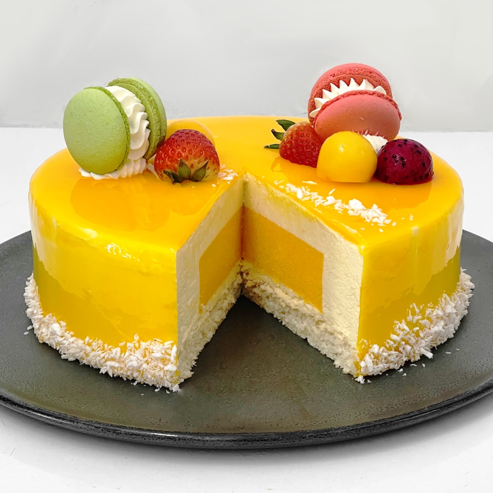 Mango mousse cake with a passionfruit filling, beautifully decorated with fresh strawberries and colorful macarons on top. The cake has a smooth, glossy finish, and the vibrant fruits add a refreshing touch.