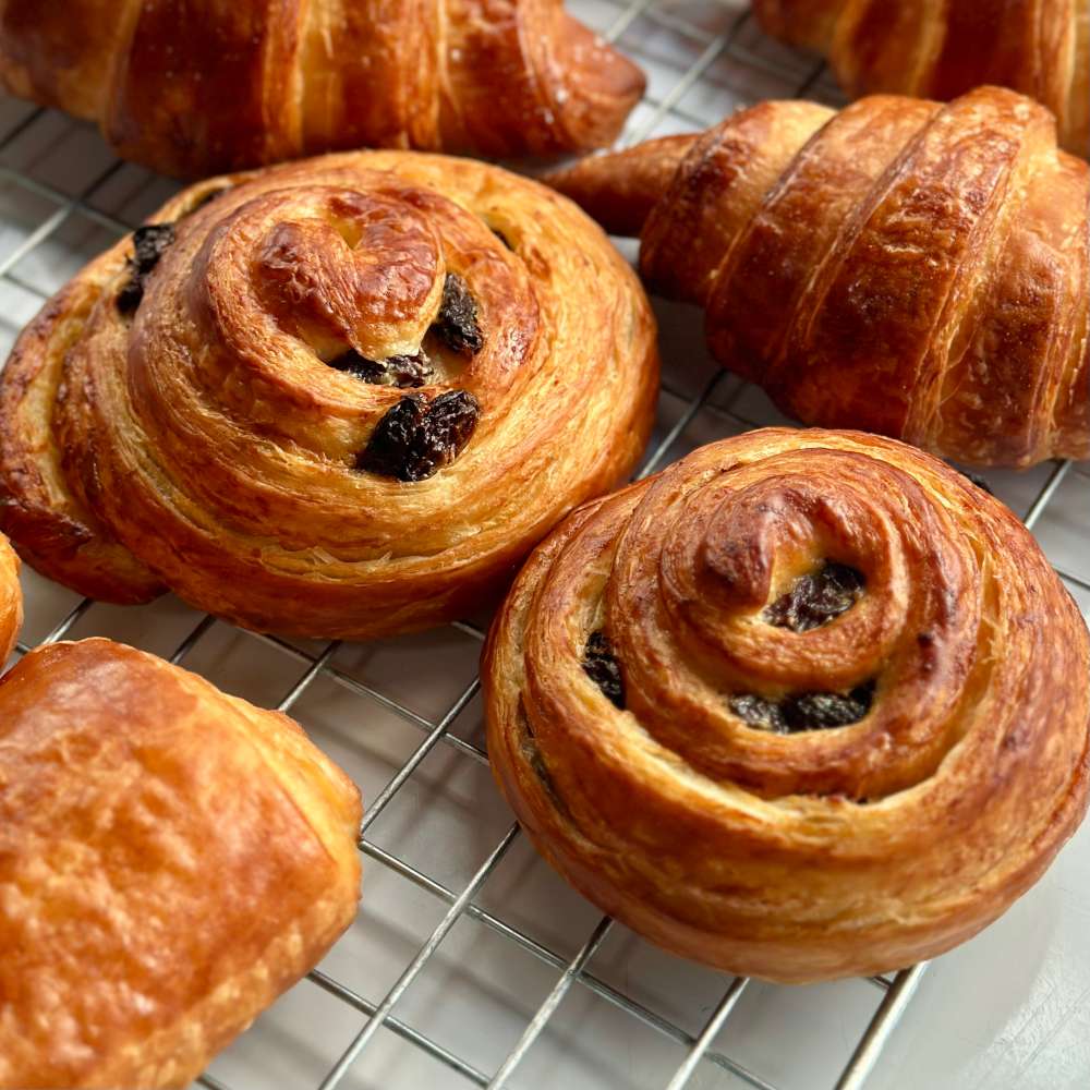 Pain au raisin, showing golden, flaky layers of croissant dough spiraled around juicy raisins and a touch of creamy filling. The pastry has a natural, matte finish, emphasizing its crisp, buttery texture.