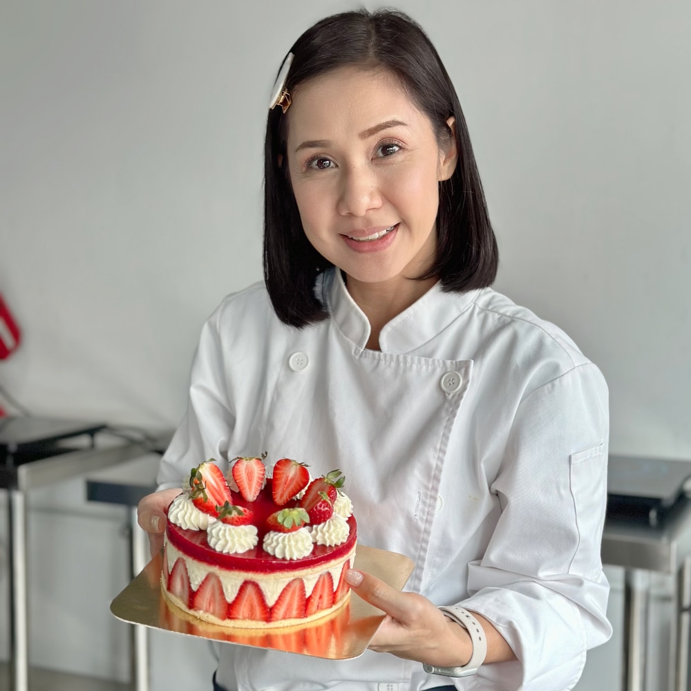 Student smiling while holding a strawberry cake, topped with fresh strawberries and a glossy finish. The student stands proudly, showcasing the beautifully crafted cake.